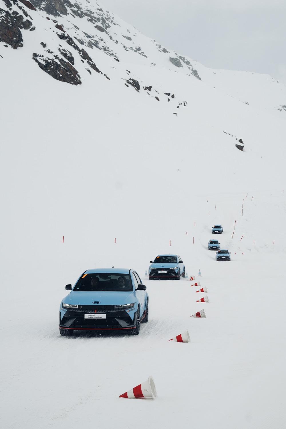 Eine Kolonne von Hyundai IONIQ 5 N Fahrzeugen auf einer schneebedeckten Bergstrecke, umgeben von verstreuten Verkehrshütchen, die ihre Fähigkeiten unter extremen Winterbedingungen demonstrieren.