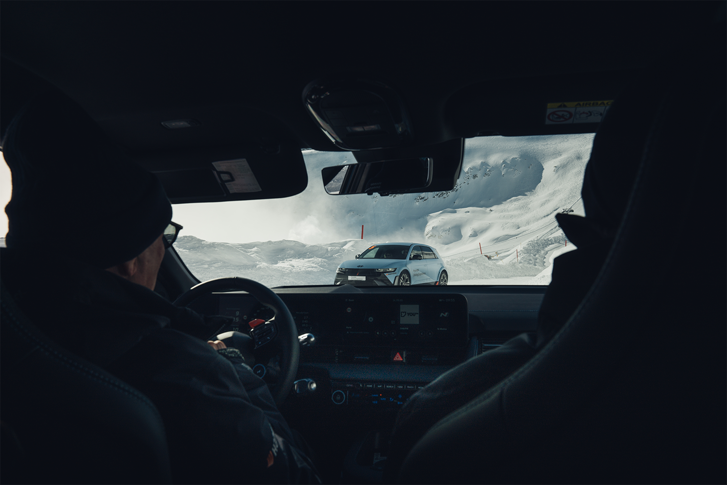 View from inside a car driving through snowy terrain, showing a Hyundai IONIQ 5 N ahead on a snowy road surrounded by mountains, framed by the car's windshield and two passengers in winter attire.