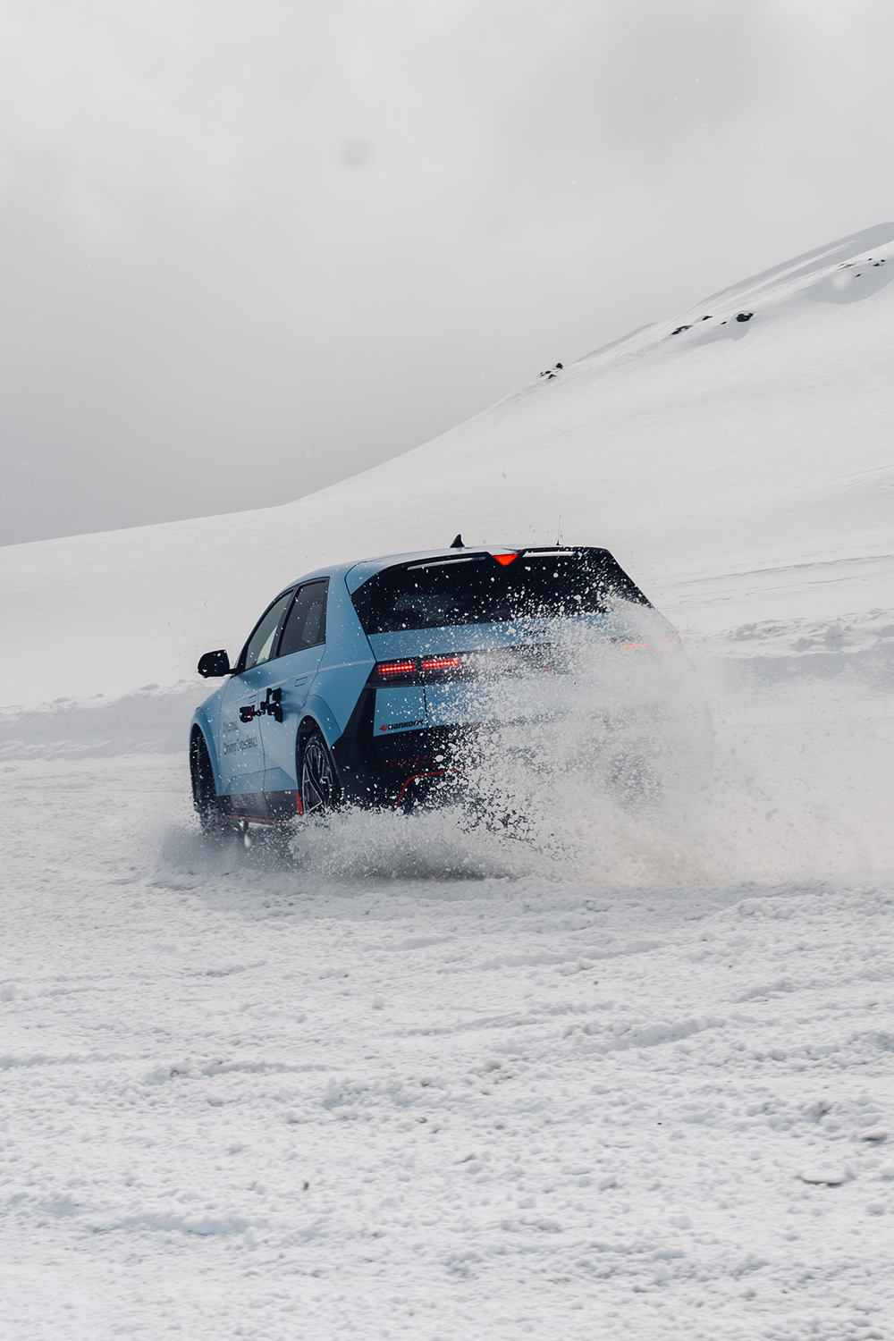 A blue Hyundai IONIQ 5 N driving through snowy terrain, with snow spraying from its tires. The vehicle's design features sharp LED lights, aerodynamic lines, and a Hyundai Driving Experience decal. The snowy mountains and clear skies form a picturesque winter backdrop.
