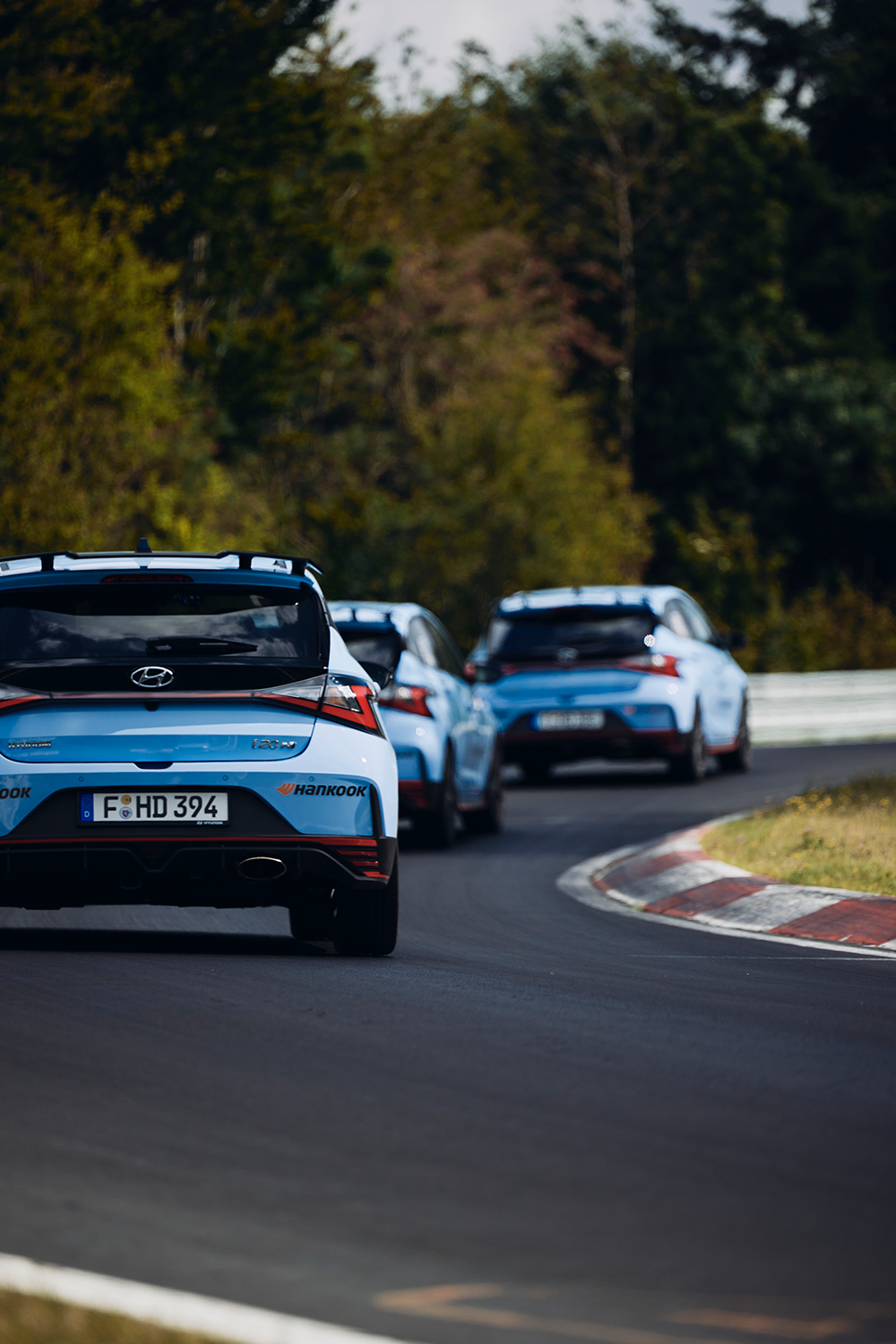 Eine Gruppe von Hyundai i20 N in Performance Blue fährt in Formation auf einer Rennstrecke, umgeben von üppigem Grün.