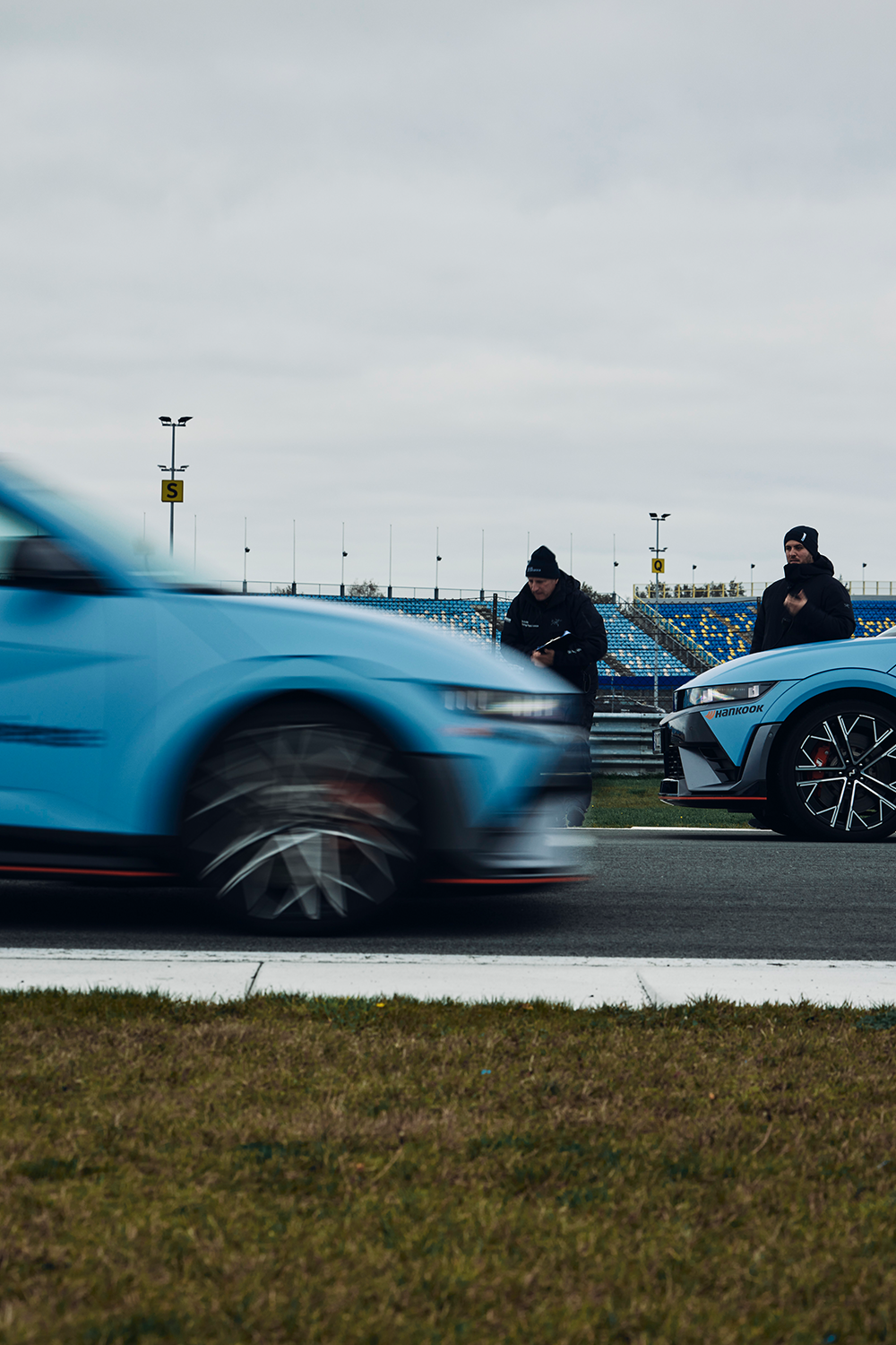 Ein Hyundai IONIQ 5 N auf einer Rennstrecke in Bewegung, wobei die verschwommene Darstellung auf hohe Geschwindigkeit hinweist. Im Hintergrund stehen zwei Personen in schwarzen Hyundai Driving Experience-Jacken neben einem weiteren IONIQ 5 N unter einem bewölkten Himmel.