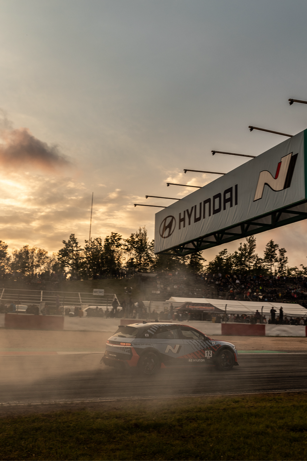 Hyundai N-Rennwagen driftet bei Sonnenuntergang auf einer Strecke, mit einem Hyundai N-Banner darüber und einer rauchigen Atmosphäre, während Zuschauer auf den Tribünen zuschauen.